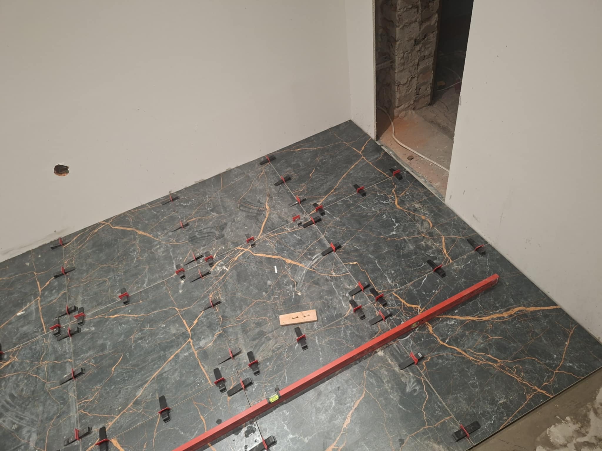 Układanie płytek podłogowych z użyciem poziomicy i klipsów poziomujących. Płytki w odcieniach szarości z wzorem imitującym marmur. Widoczny fragment ściany i otwór w niej.