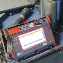 Miernik Sonel MPI-540 z podłączonymi przewodami pomiarowymi, wyświetlający wynik pomiaru rezystancji. Urządzenie stoi na wózku warsztatowym. Widoczne złącza USB i gniazda pomiarowe.