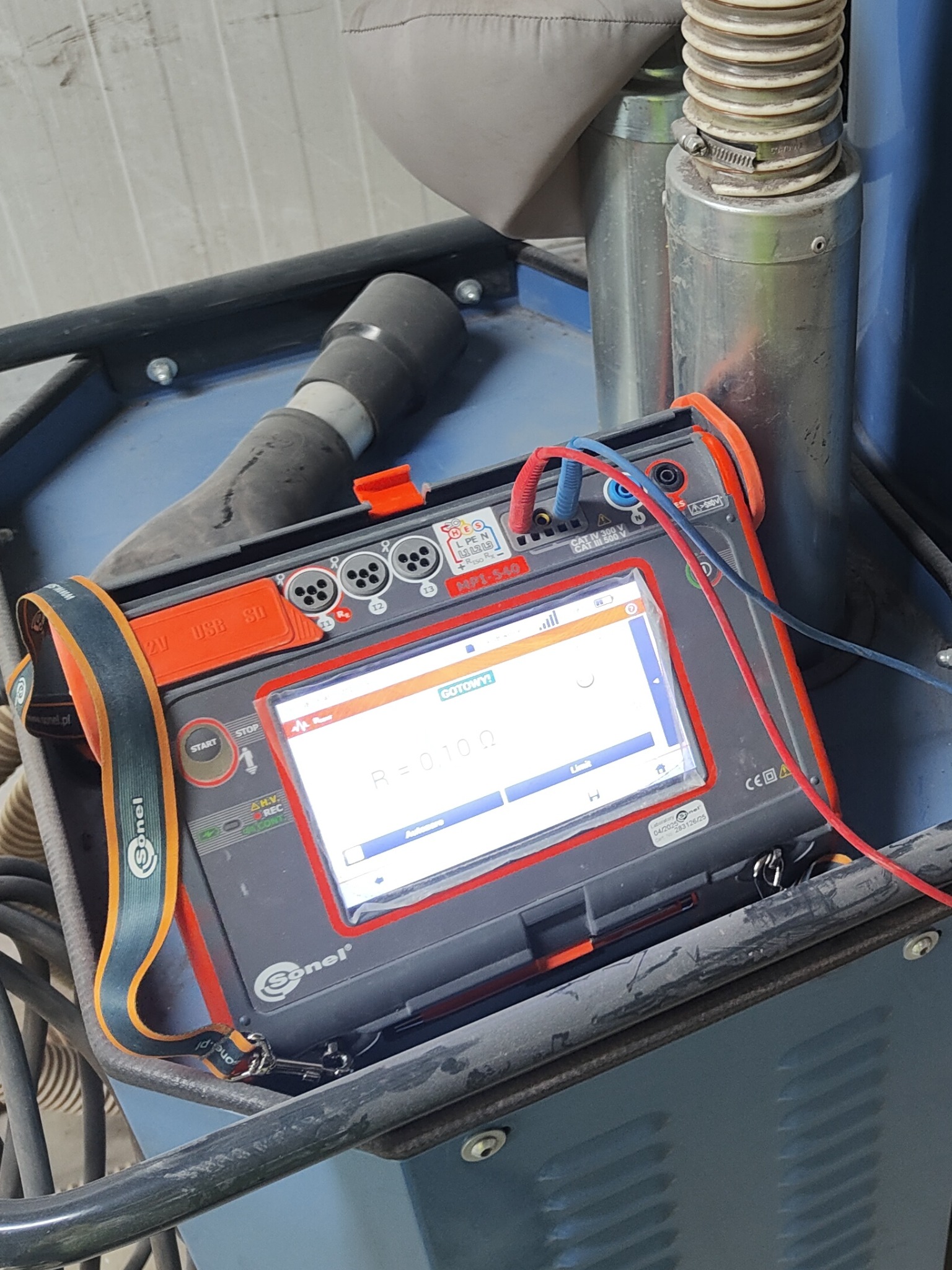 Miernik Sonel MPI-540 z podłączonymi przewodami pomiarowymi, wyświetlający wynik pomiaru rezystancji. Urządzenie stoi na wózku warsztatowym. Widoczne złącza USB i gniazda pomiarowe.