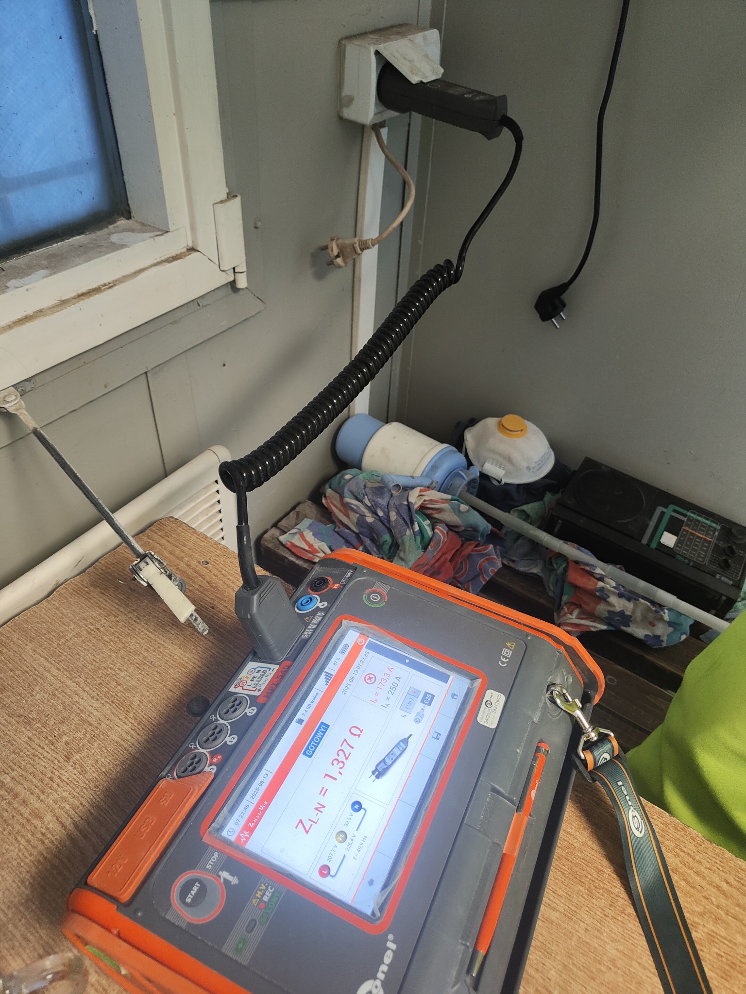 Miernik elektryczny Anel MPI-540 pokazuje impedancję pętli zwarcia ZL-N 1,327 Ω. W tle gniazdko elektryczne, okno i przedmioty leżące na stole.