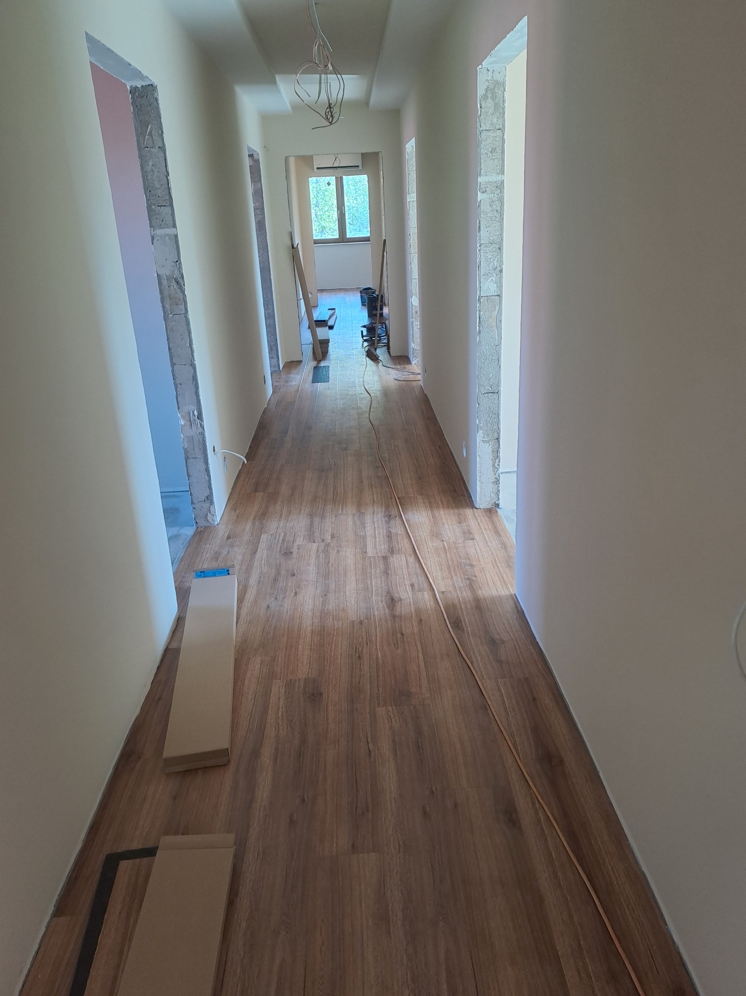 Korytarz po montażu paneli podłogowych z widocznymi fragmentami nieułożonych desek. Widoczne kable i przygotowane otwory drzwiowe.