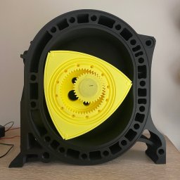 Mobilna Kraina EDEN Witold Perucki - Czarno-żółty model silnika Wankla, wydrukowany w 3D, z widocznymi detalami kół zębatych i obudowy, stojący na drewnianym blacie. Model prezentowany w neutralnym oświetleniu.