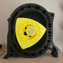 Czarno-żółty model silnika Wankla, wydrukowany w 3D, z widocznymi detalami kół zębatych i obudowy, stojący na drewnianym blacie. Model prezentowany w neutralnym oświetleniu.