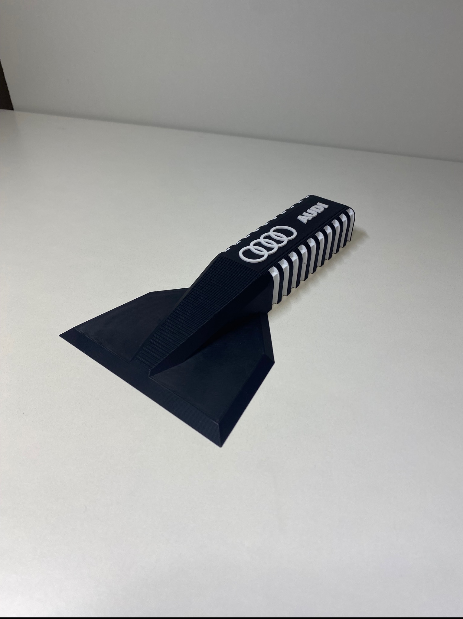 Czarna, drukowana w 3D skrobaczka do szyb z logo Audi, leżąca na białym tle. Widoczne charakterystyczne warstwy druku 3D.