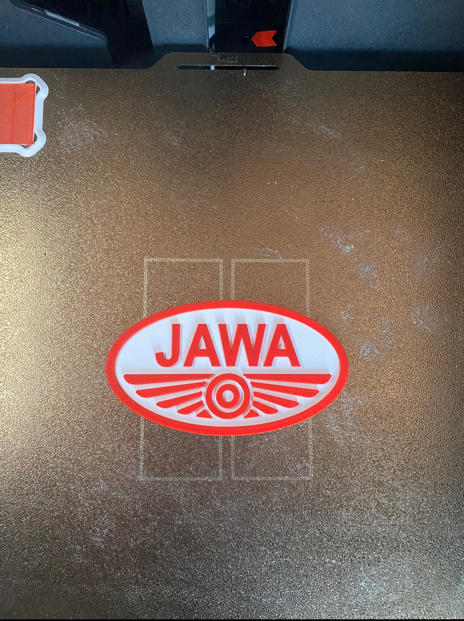 Logo JAWA, wydrukowane w 3D z czerwono-białego filamentu, leży na teksturowanej powierzchni roboczej drukarki 3D. Widoczne ślady użytkowania na stole.