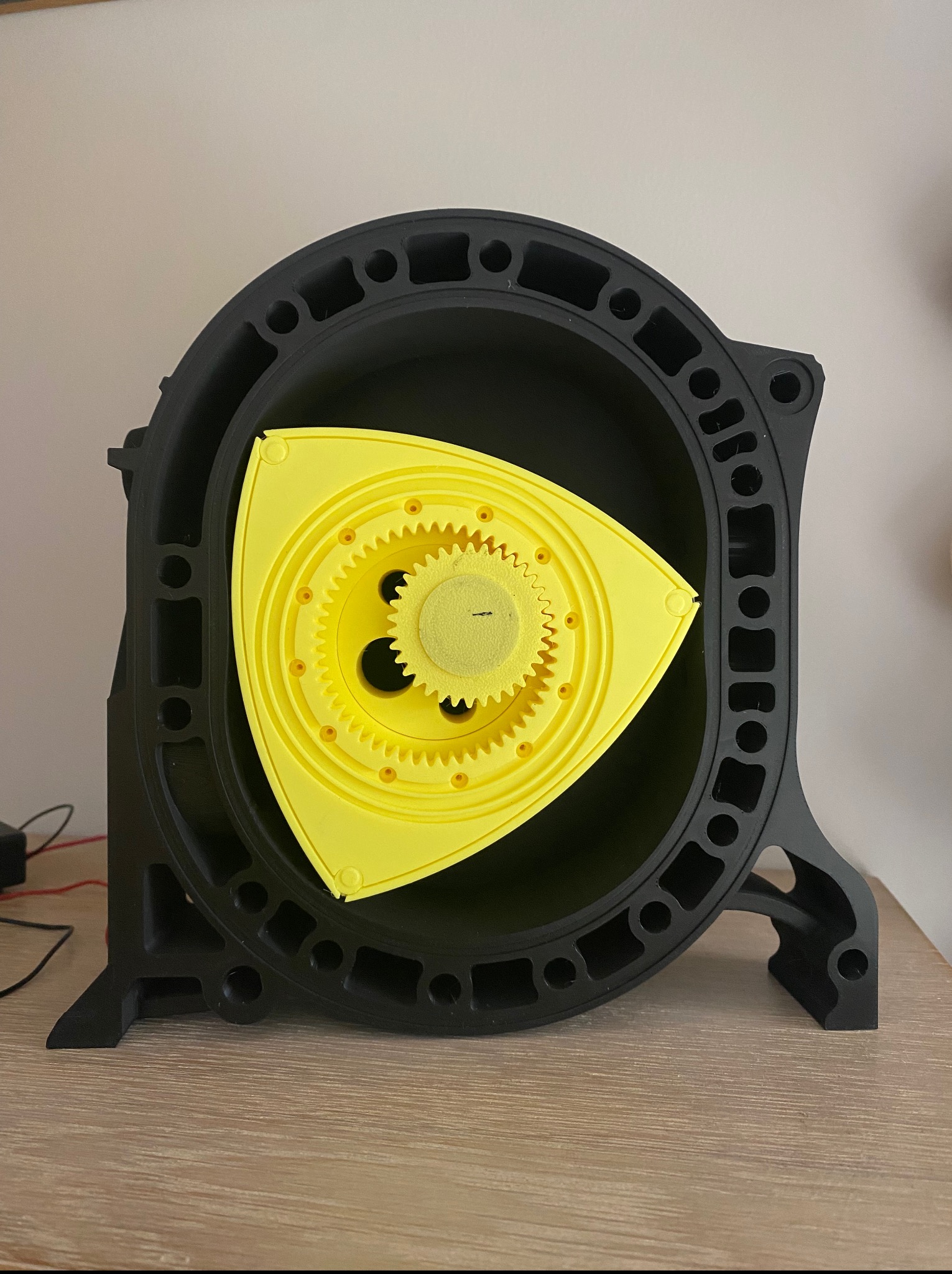 Czarno-żółty model silnika Wankla, wydrukowany w 3D, z widocznymi detalami kół zębatych i obudowy, stojący na drewnianym blacie. Model prezentowany w neutralnym oświetleniu.