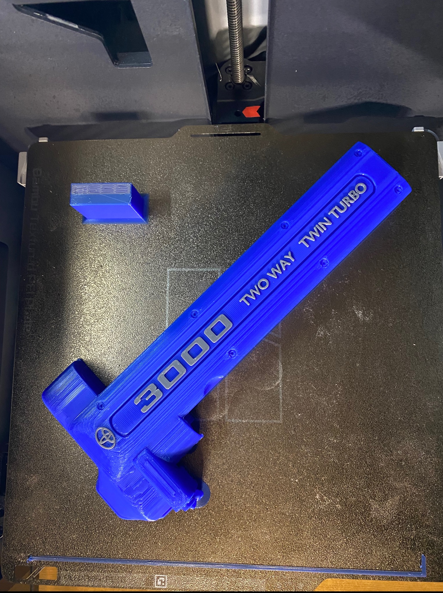 Niebieski element z napisem '3000 TWO WAY TWIN TURBO' wydrukowany w 3D leży na platformie drukarki 3D, obok widoczny mały, niebieski sześcian.