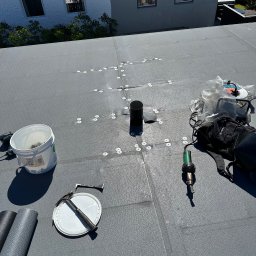 Piotr Nik - Szary dach płaski z narzędziami dekarskimi i elementami mocującymi, widok z góry. W tle fragment budynku mieszkalnego.