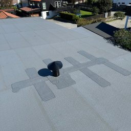 Piotr Nik - Szary dach z membraną i kominkiem wentylacyjnym na tle zabudowy jednorodzinnej w słoneczny dzień. Widoczne łączenia membrany.