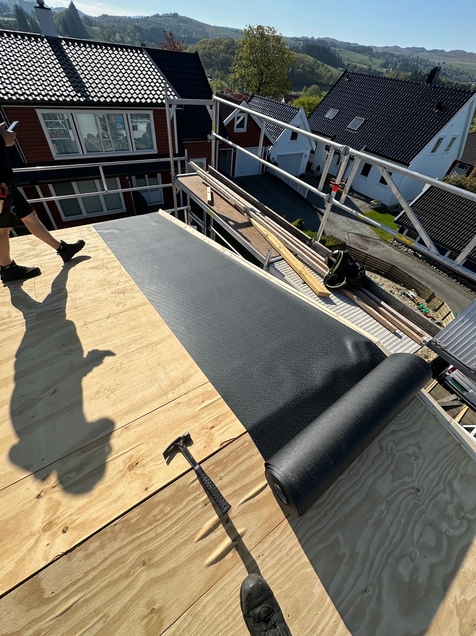 Montaż papy na dachu z desek. Widoczny młotek, rolka papy i fragment sylwetki dekarza. W tle domy i wzgórza w słoneczny dzień.