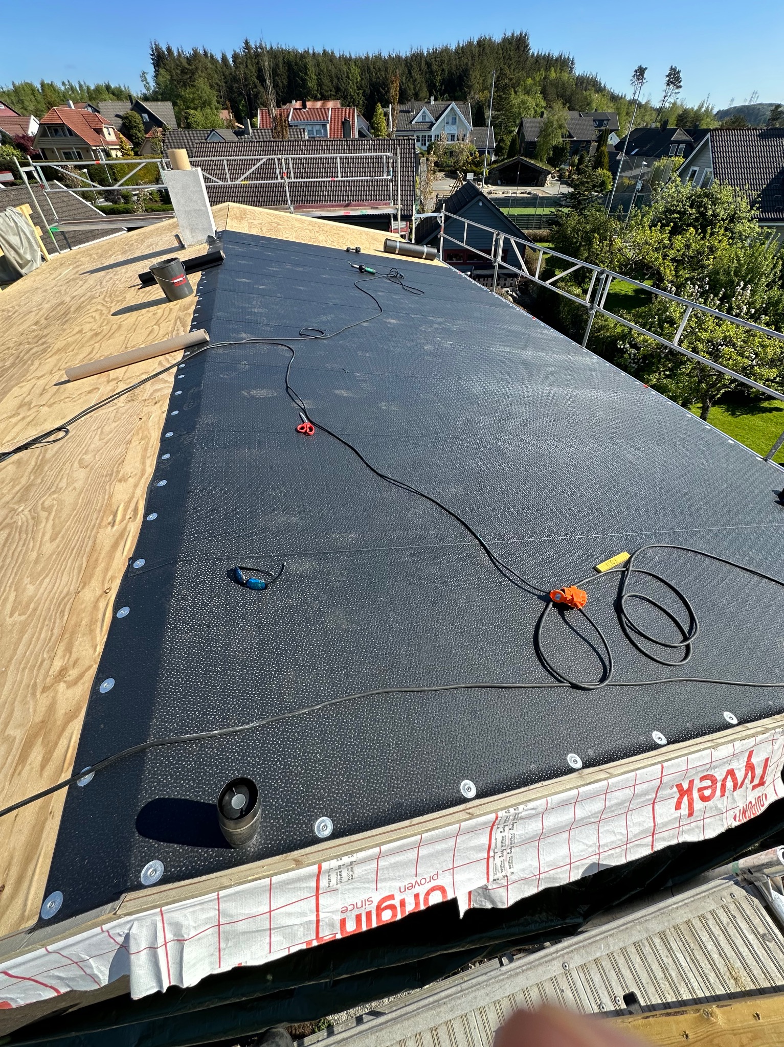 Montaż czarnej membrany dachowej na drewnianej konstrukcji, widoczne narzędzia i elementy mocujące, w tle zabudowa jednorodzinna i zieleń. Praca w toku, jasne oświetlenie.