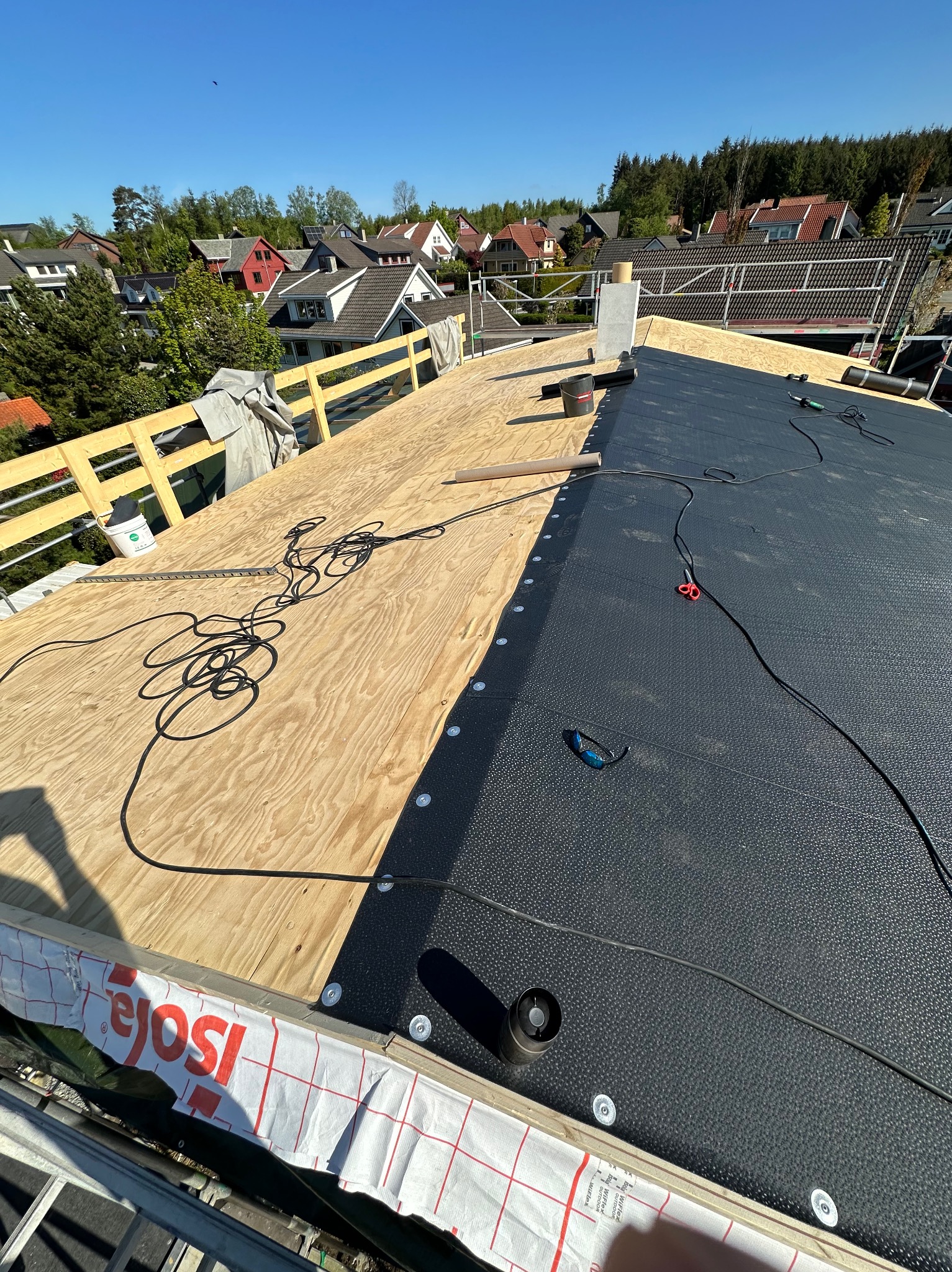 Dach w trakcie krycia: część z płyt OSB, część z czarną membraną. Widoczne narzędzia, kable i elementy mocujące. W tle zabudowa jednorodzinna i zieleń.