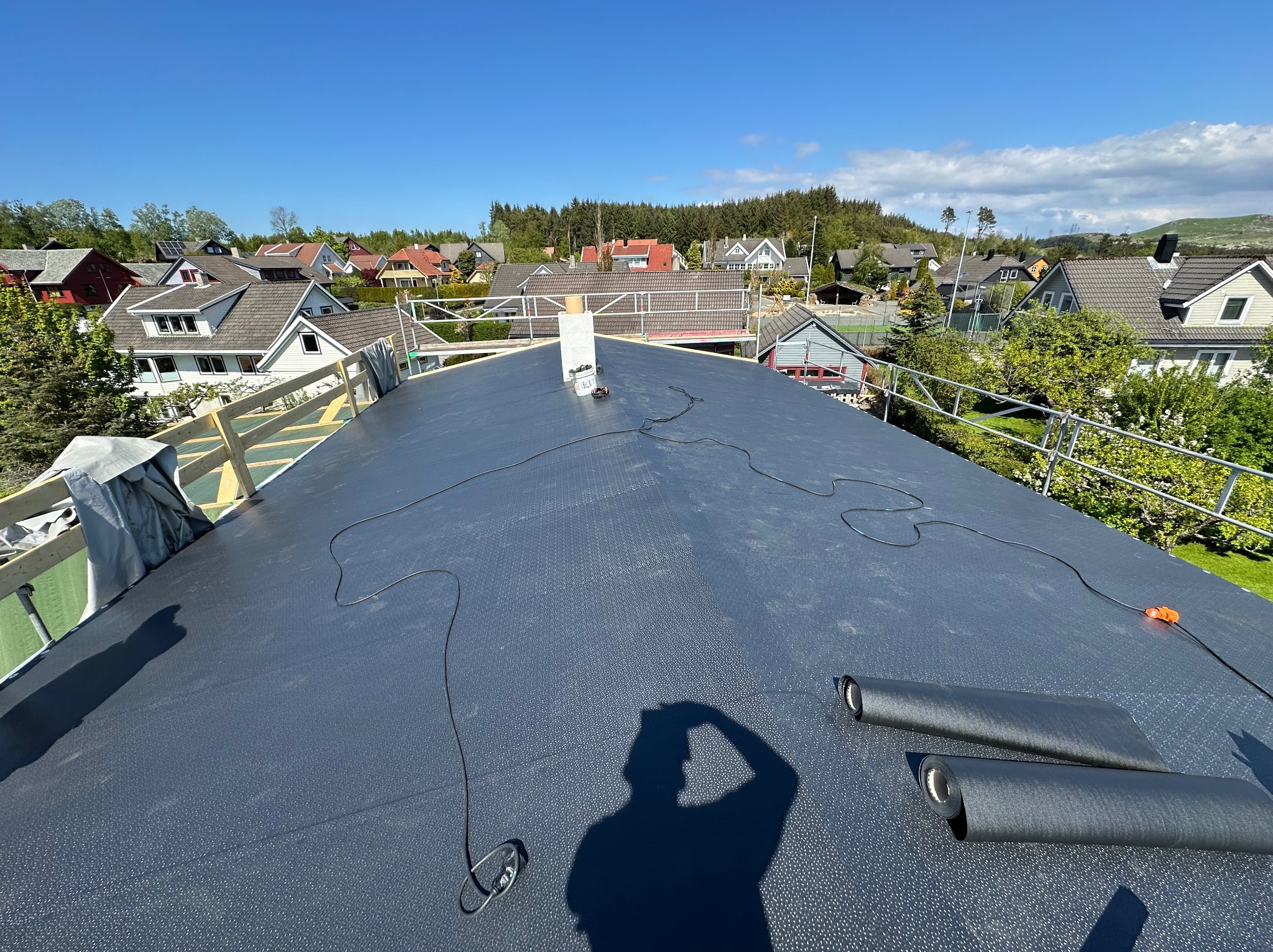 Czarny, płaski dach pokryty papą termozgrzewalną w trakcie prac dekarskich. W tle zabudowa jednorodzinna i błękitne niebo. Widoczny cień osoby wykonującej zdjęcie.