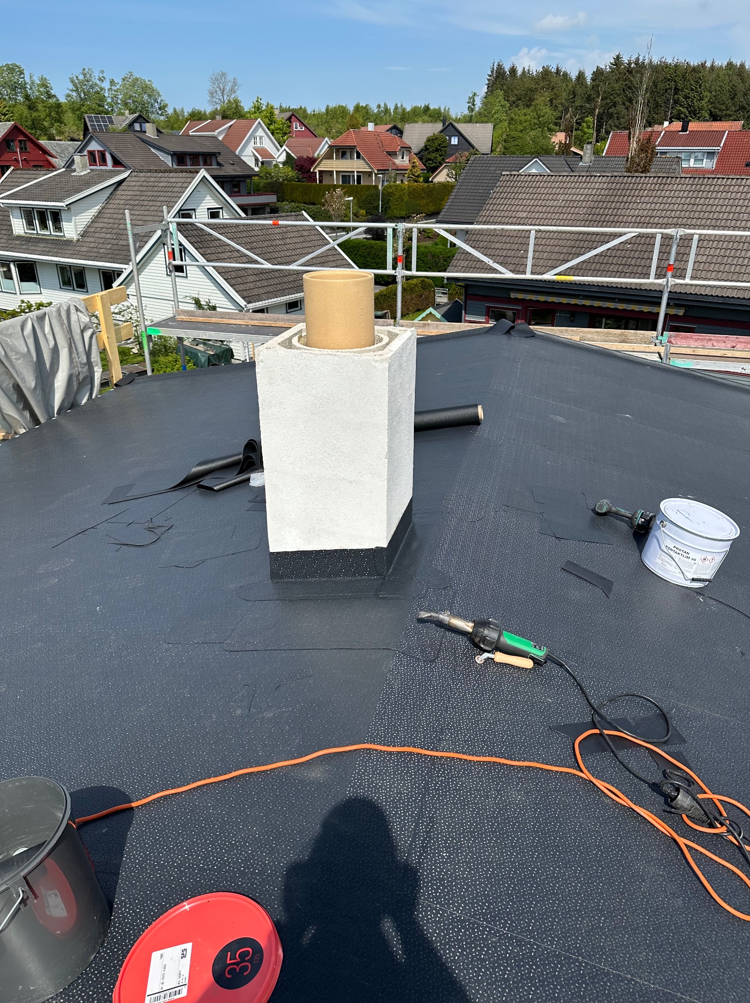 Czarny dach pokryty papą termozgrzewalną w trakcie prac dekarskich. Widoczny komin, narzędzia dekarskie i okoliczne domy w tle. Słoneczny dzień.