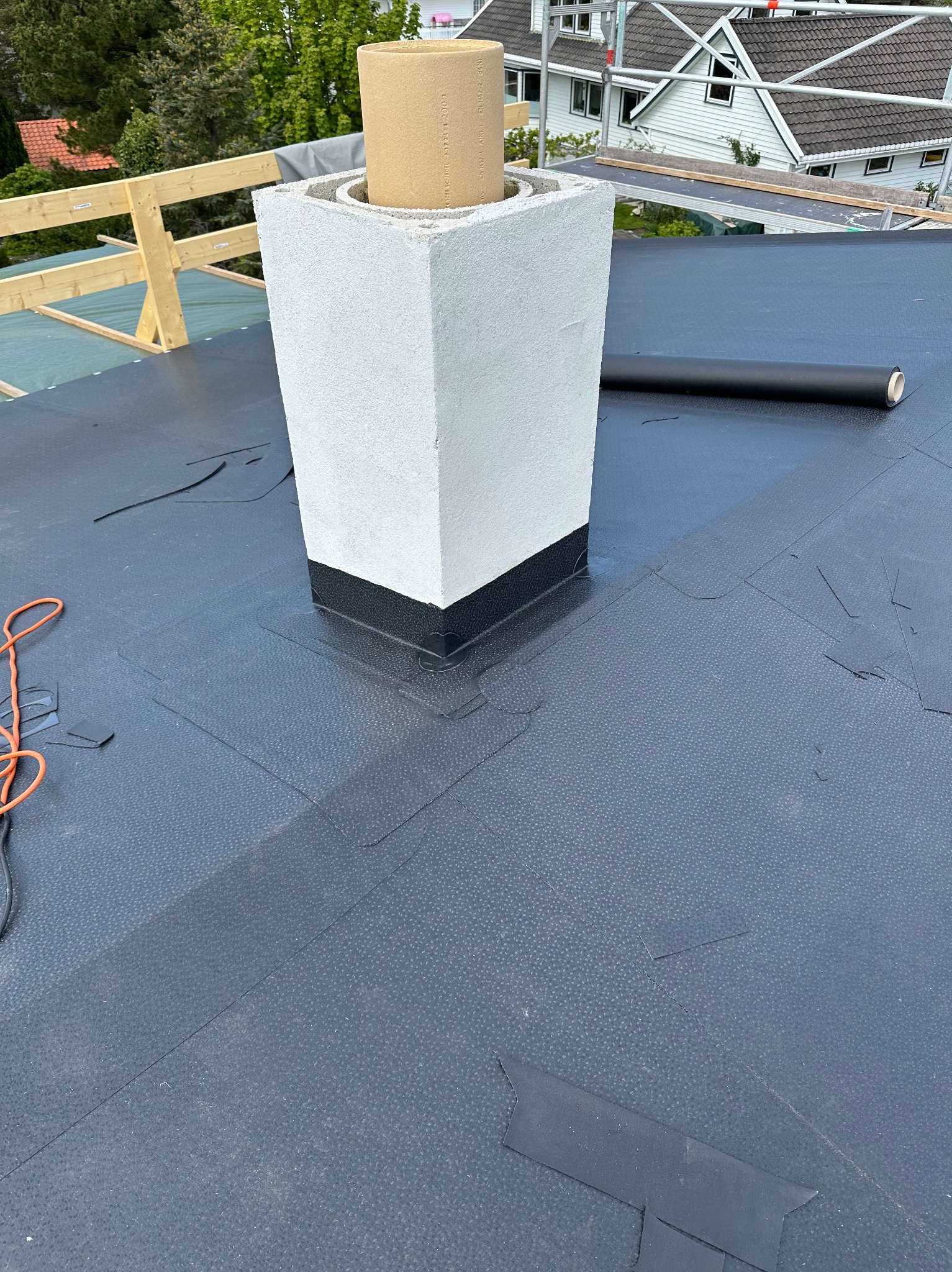 Czarny dach z papy z kominem w trakcie budowy. Widoczny fragment zwiniętej rolki papy i kabel elektryczny. W tle biały dom z szarym dachem.