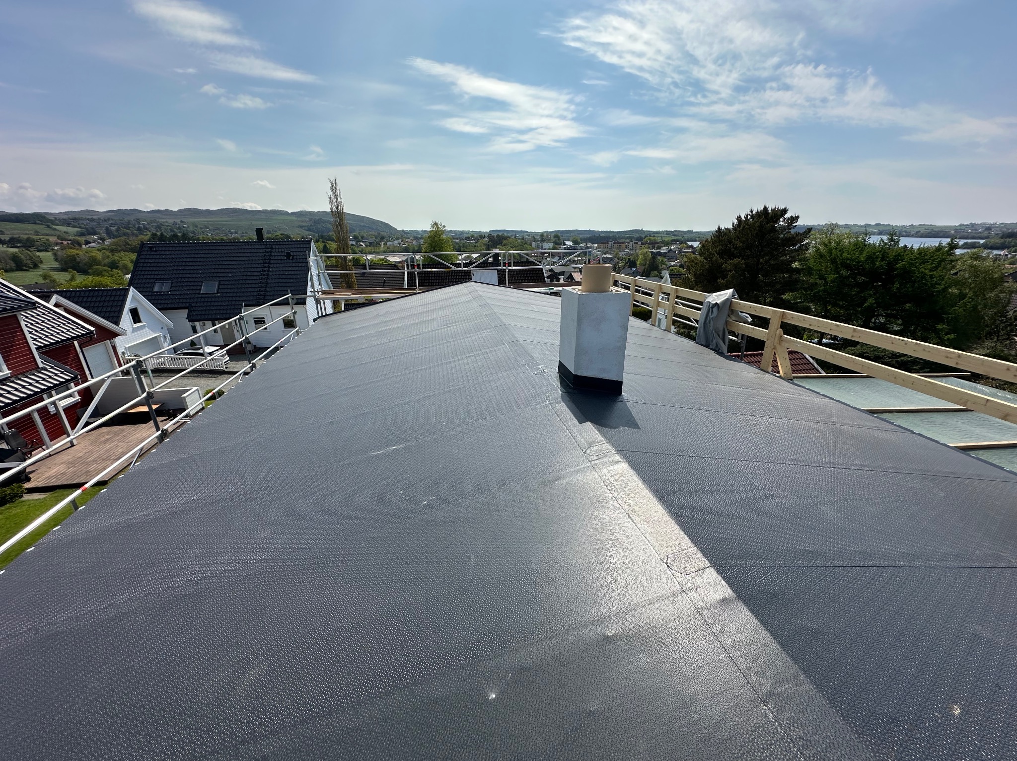 Czarny, płaski dach z kominem w trakcie prac dekarskich, zabezpieczony barierkami. W tle zabudowa mieszkalna i zielony krajobraz pod błękitnym niebem.