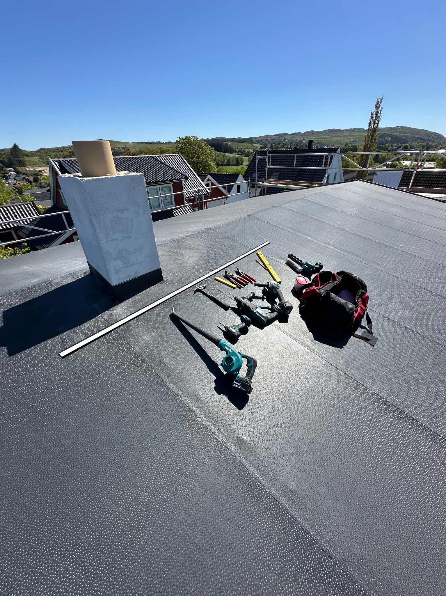 Szary dach z papy w Bielsko-Białej, narzędzia dekarskie i komin. Widok na okoliczne domy i wzgórza w słoneczny dzień. Praca dekarska w toku.