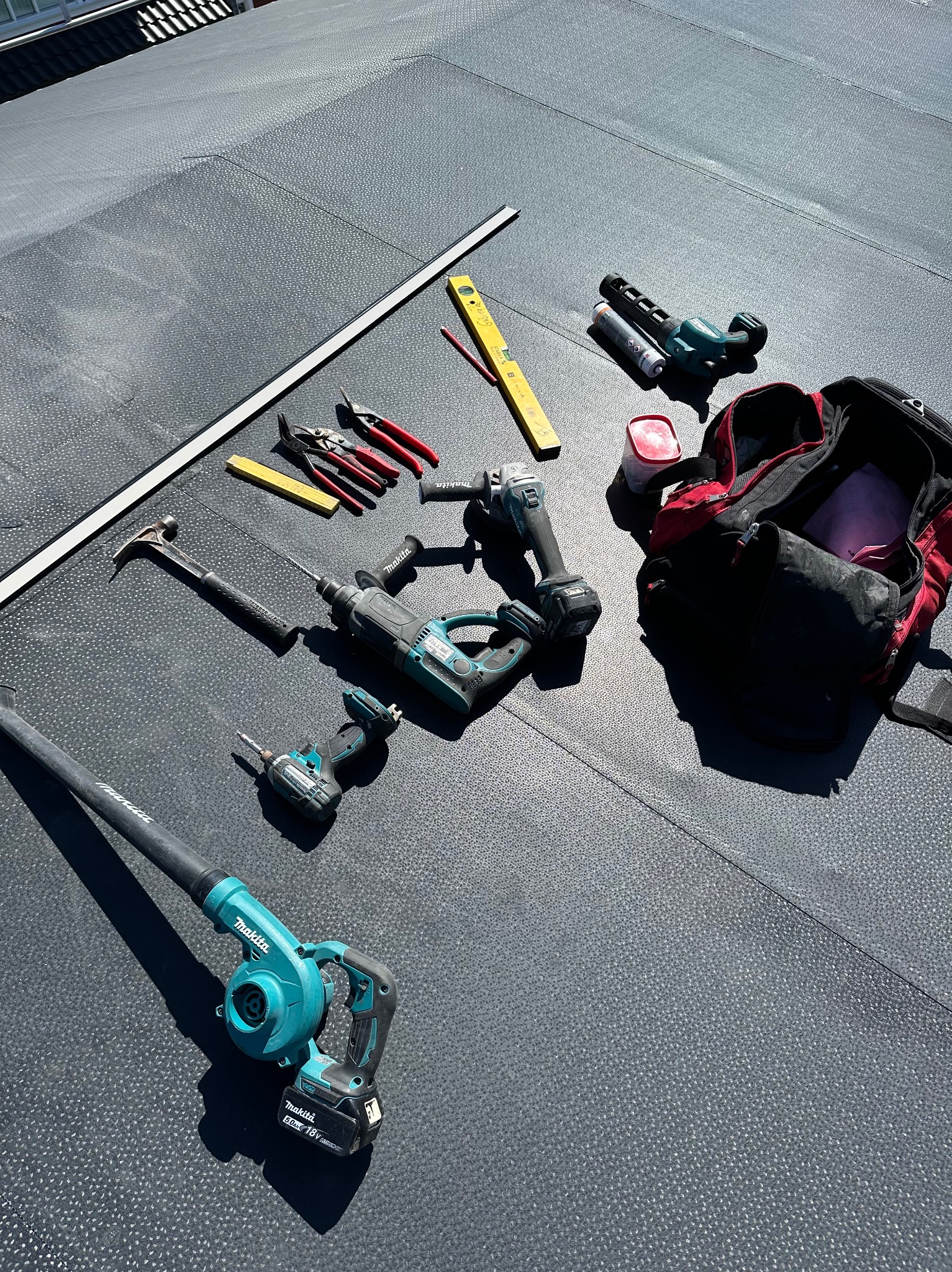 Narzędzia dekarskie Makita rozłożone na czarnym, płaskim dachu z papy, obok czerwono-czarnego plecaka narzędziowego. Widoczny poziomica, młotek i wkrętarka.