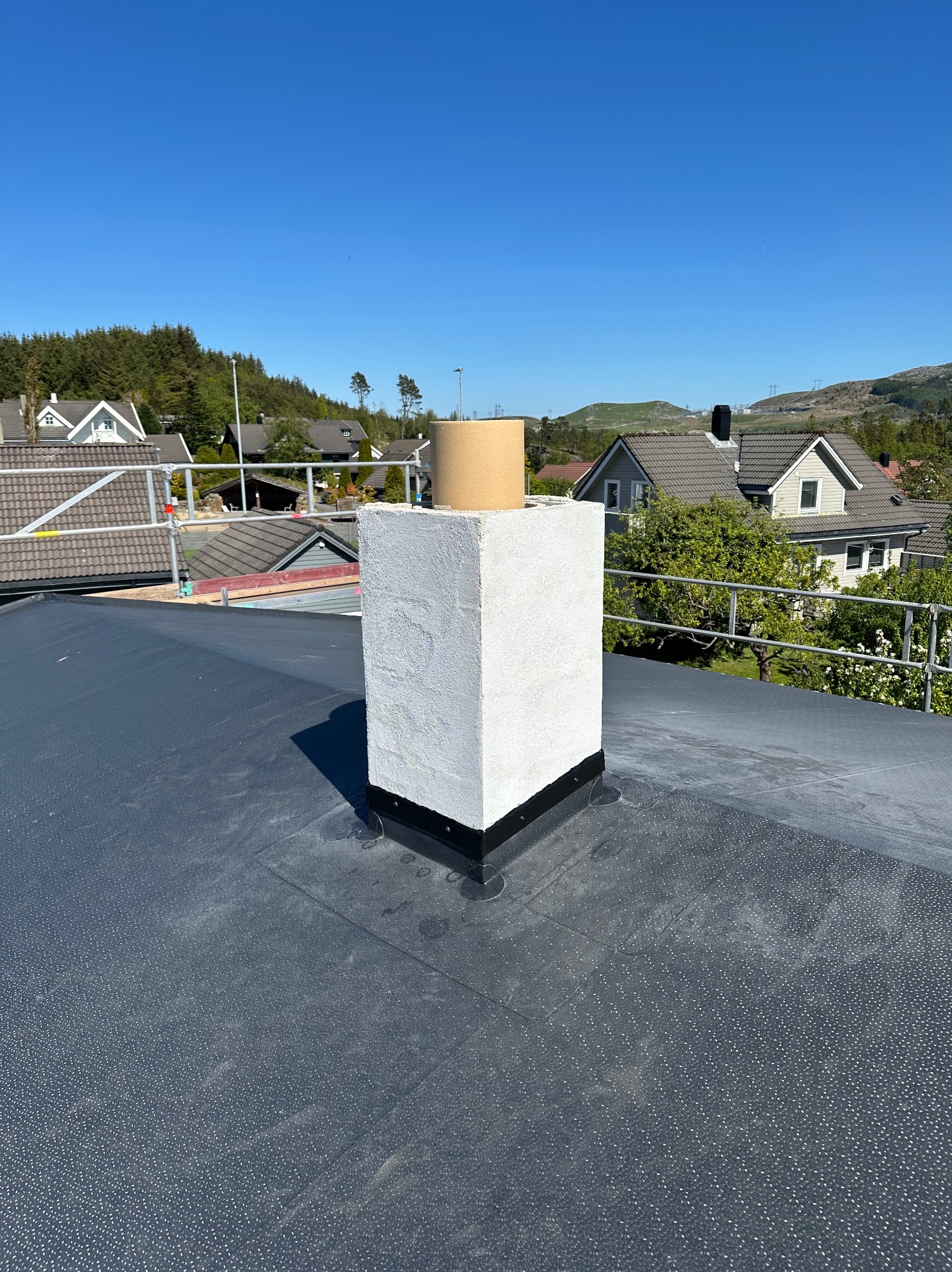 Wykończony komin na dachu pokrytym papą, z widocznymi elementami uszczelniającymi. W tle zabudowa jednorodzinna i zieleń w słoneczny dzień.