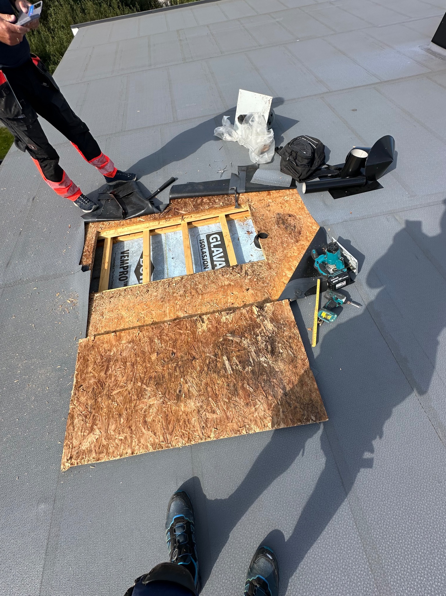 Fragment dachu z odsłoniętą izolacją, narzędzia dekarskie i fragmenty desek. Widoczne elementy instalacji i ekipa dekarska w trakcie pracy.