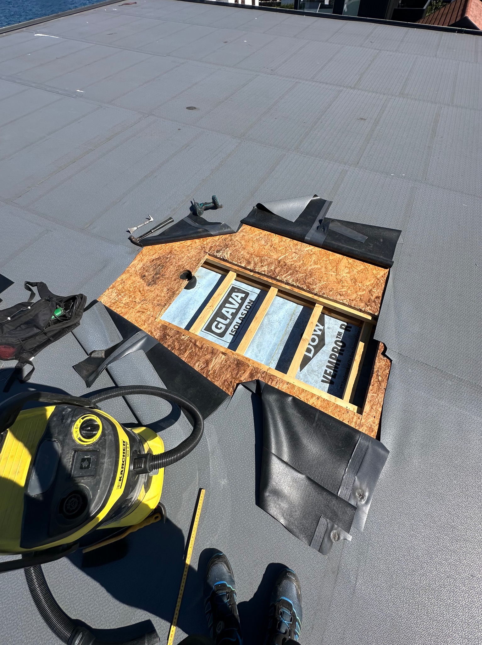 Naprawa dachu z papy: odsłonięta warstwa izolacyjna Glava i Dow Vepro, narzędzia, odkurzacz Karcher, buty dekarza. Widok z góry na płaski dach podczas prac.