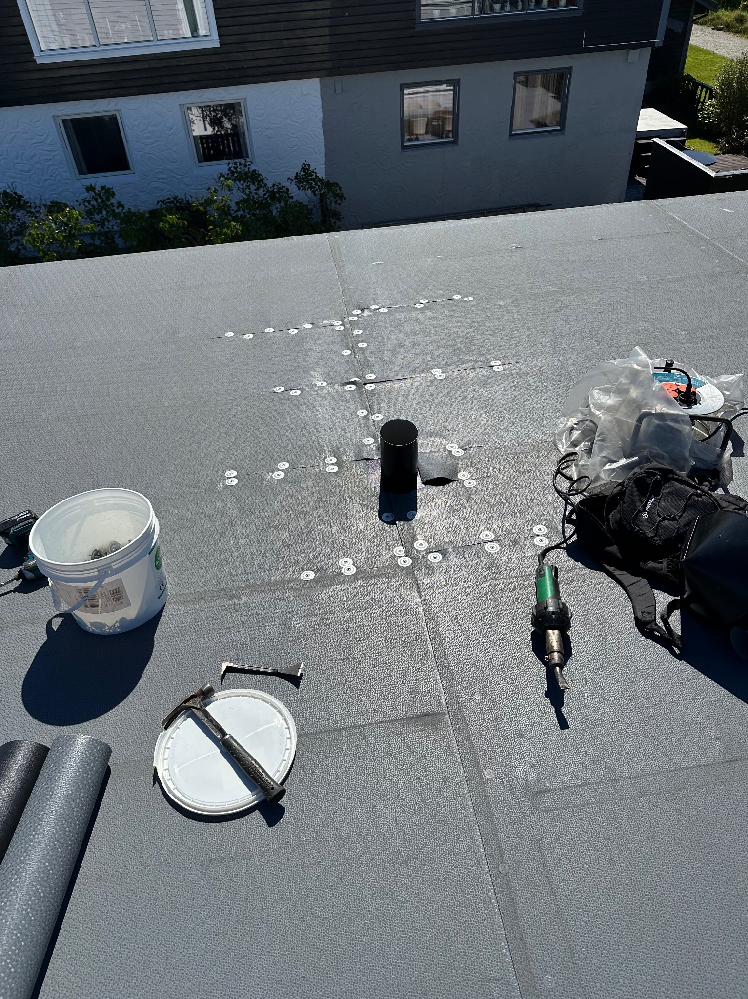 Szary dach płaski z narzędziami dekarskimi i elementami mocującymi, widok z góry. W tle fragment budynku mieszkalnego.