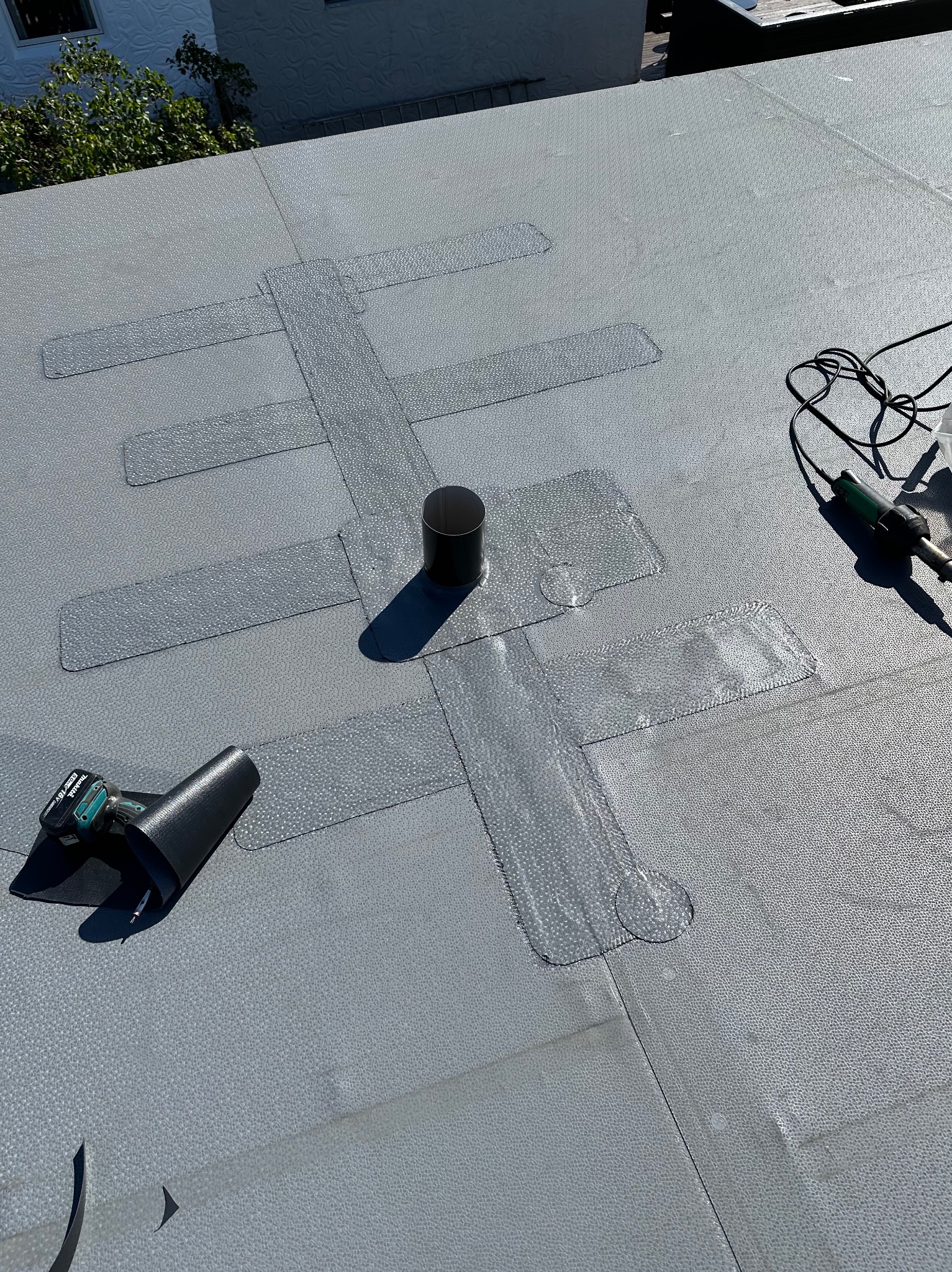 Szary dach płaski z uszczelnieniami wokół komina i narzędziami dekarskimi. Widoczne zgrzewy papy i narzędzia do ich wykonania. Detal dachu po naprawie.