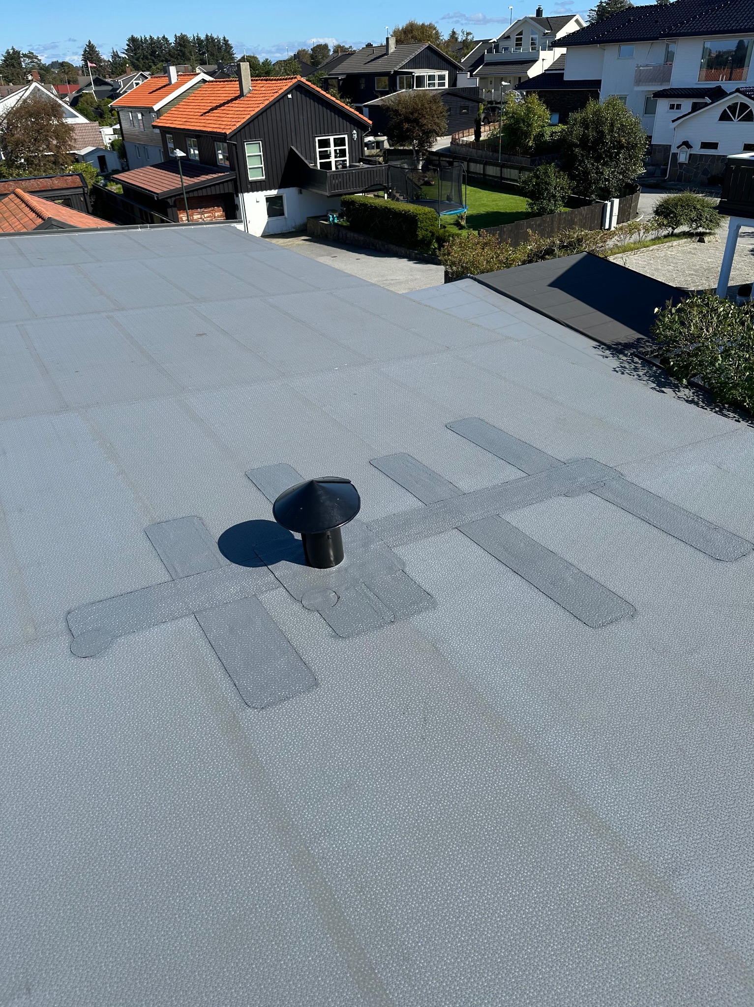 Szary dach z membraną i kominkiem wentylacyjnym na tle zabudowy jednorodzinnej w słoneczny dzień. Widoczne łączenia membrany.