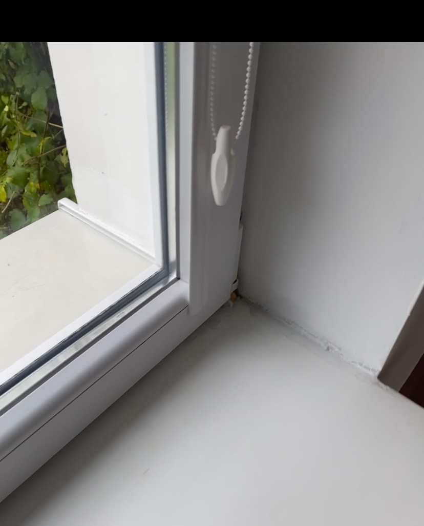 Białe okno PCV z roletą zwiniętą u góry, widoczny parapet i fragment ściany. Plamy zabrudzeń w rogu okna i na parapecie.