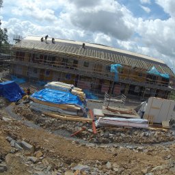 Holhauser - Wznoszenie dwukondygnacyjnego budynku z drewnianą konstrukcją dachu. Na rusztowaniach pracownicy, teren budowy z materiałami i ziemią. Szerszy kąt widzenia.