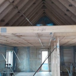 Holhauser - Wnętrze domu w trakcie budowy: widoczne drewniane elementy konstrukcyjne dachu i stropu, ściany w folii ochronnej, podpory oraz instalacje. Surowy stan wykończenia.