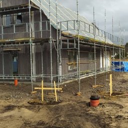 Holhauser - Dwukondygnacyjny dom w trakcie budowy, otoczony rusztowaniami. Widoczne elementy instalacji zewnętrznej i teren przygotowany pod dalsze prace.