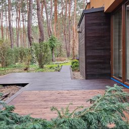 Holhauser - Ciemnobrązowy, drewniany dom z tarasem w otoczeniu drzew. Taras wykonany z desek w dwóch odcieniach brązu. Widoczne zabezpieczenia okien folią.