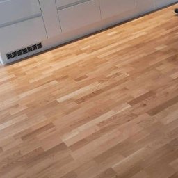 Holhauser - Jasna, drewniana podłoga ułożona w jodełkę w nowoczesnej kuchni z białymi szafkami i ciemną mozaiką nad blatem. Widoczny fragment osoby w prawym górnym rogu.