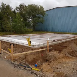 Zbrojenie fundamentów pod budowę, widok z góry. Stalowa siatka zbrojeniowa na białej izolacji, drewniane szalunki. W tle budynek przemysłowy i drzewa.