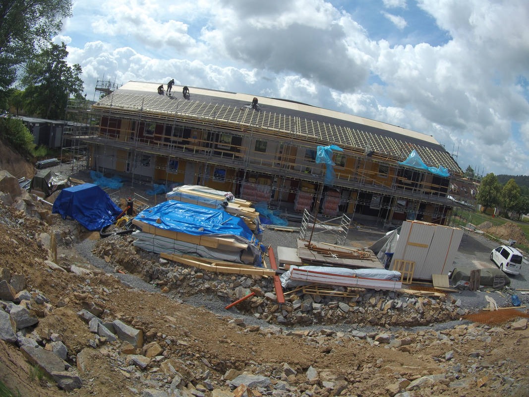 Wznoszenie dwukondygnacyjnego budynku z drewnianą konstrukcją dachu. Na rusztowaniach pracownicy, teren budowy z materiałami i ziemią. Szerszy kąt widzenia.