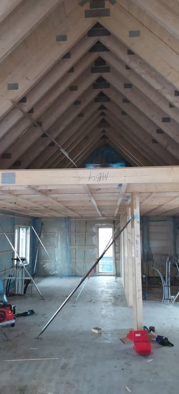 Wnętrze domu w trakcie budowy: widoczne drewniane elementy konstrukcyjne dachu i stropu, ściany w folii ochronnej, podpory oraz instalacje. Surowy stan wykończenia.