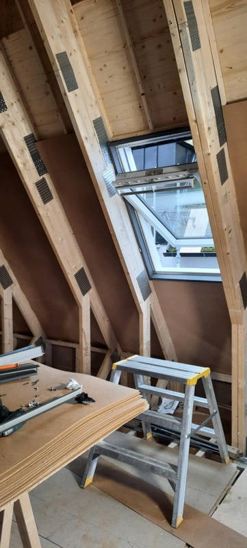 Poddasze w trakcie prac wykończeniowych: widoczne drewniane belki, okno dachowe i stos płyt. Metalowa drabina i narzędzia sugerują trwający montaż.