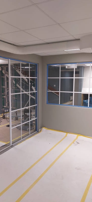 Wnętrze w trakcie prac wykończeniowych: zabezpieczona podłoga, okna i przesuwne drzwi z białymi szprosami, widoczne rusztowania i sprzęt budowlany.