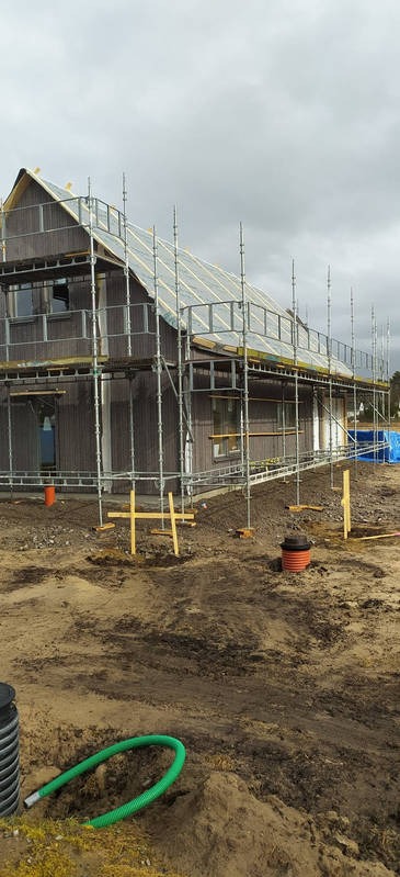 Dwukondygnacyjny dom w trakcie budowy, otoczony rusztowaniami. Widoczne elementy instalacji zewnętrznej i teren przygotowany pod dalsze prace.