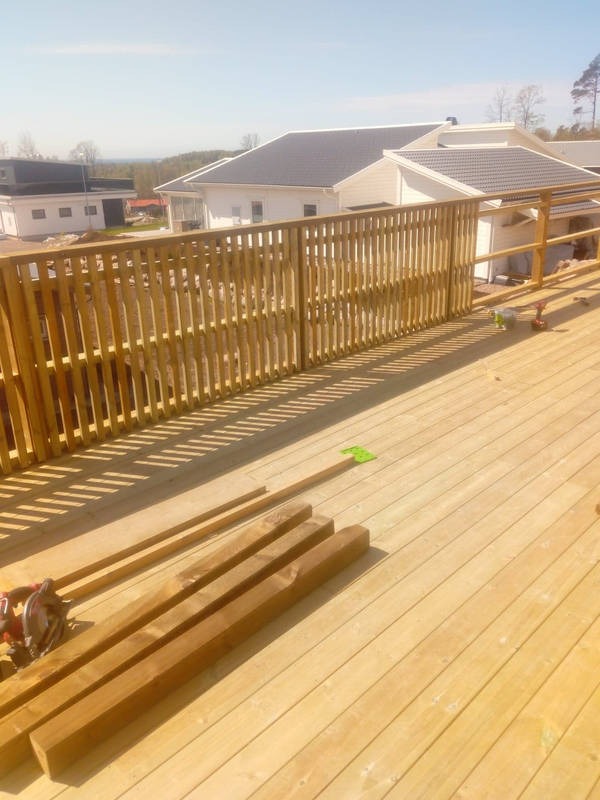 Jasna, drewniana balustrada tarasu w trakcie budowy, z widocznymi deskami, narzędziami i domem w tle. Słoneczny dzień, świeże drewno.