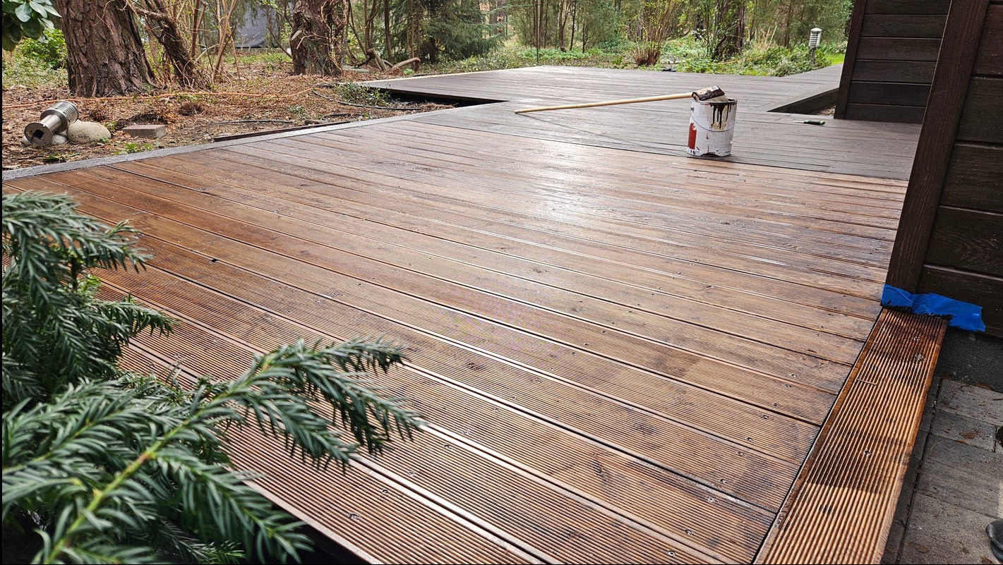 Impregnowana drewniana podłoga tarasu z widoczną puszką farby i pędzlem, podkreślająca detale wykończenia i estetykę. Częściowo pomalowana podłoga.