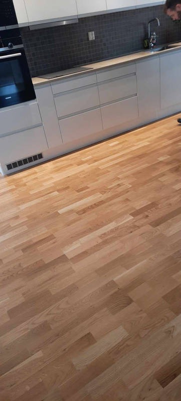 Jasna, drewniana podłoga ułożona w jodełkę w nowoczesnej kuchni z białymi szafkami i ciemną mozaiką nad blatem. Widoczny fragment osoby w prawym górnym rogu.