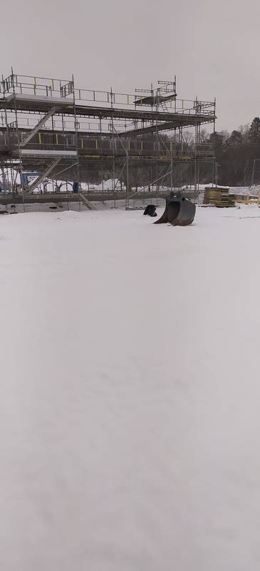 Stalowe rusztowanie na zasypanym śniegiem placu budowy, z fragmentem łyżki koparki i paletami w tle, w pochmurny dzień. Widoczne drzewa w oddali.