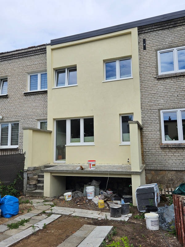 Żółta, nowa elewacja dobudowanej części domu z oknami, kontrastująca z ceglanym budynkiem obok. Widoczne ślady prac wykończeniowych i narzędzia.