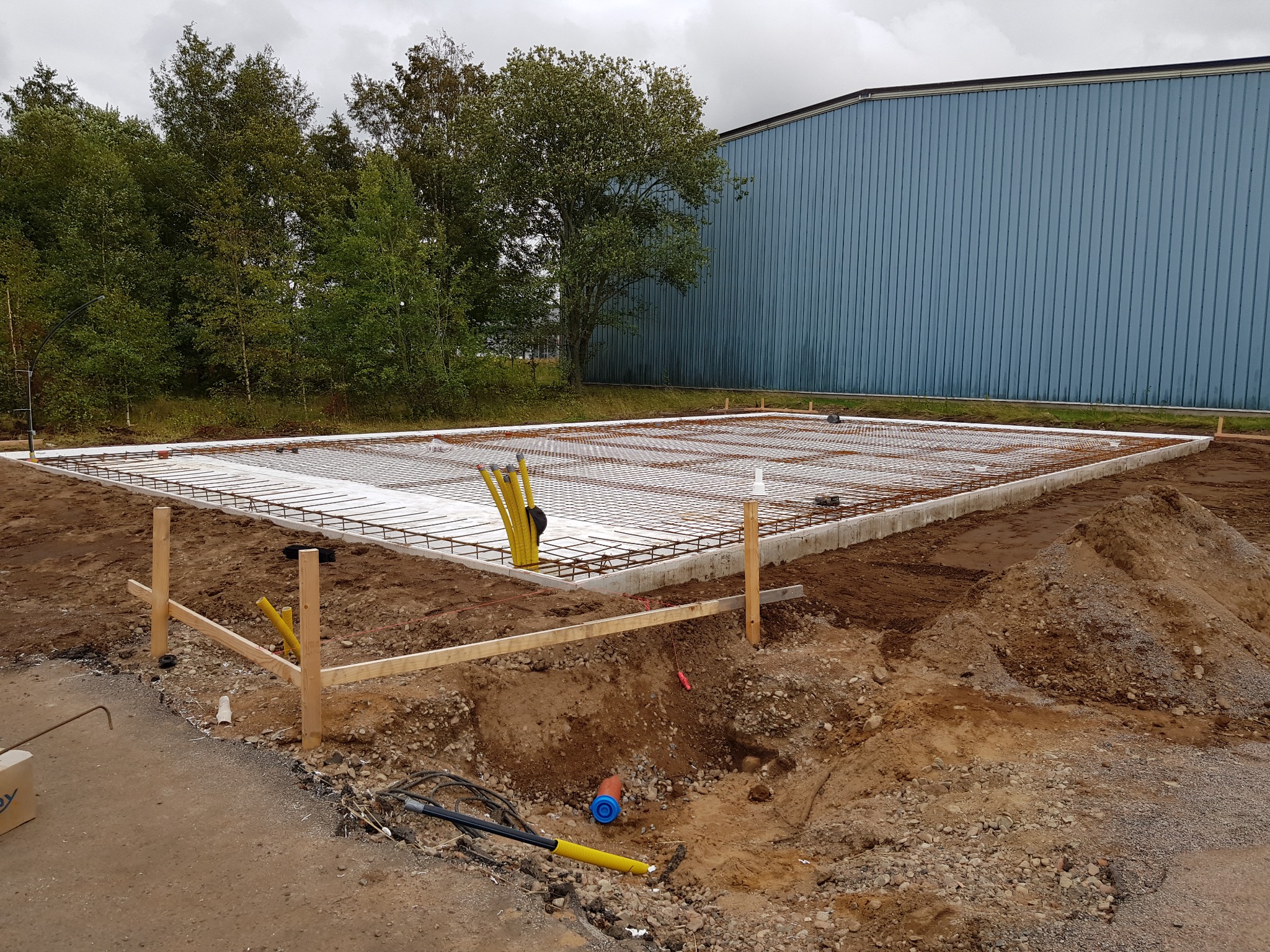 Zbrojenie fundamentów pod budowę, widok z góry. Stalowa siatka zbrojeniowa na białej izolacji, drewniane szalunki. W tle budynek przemysłowy i drzewa.