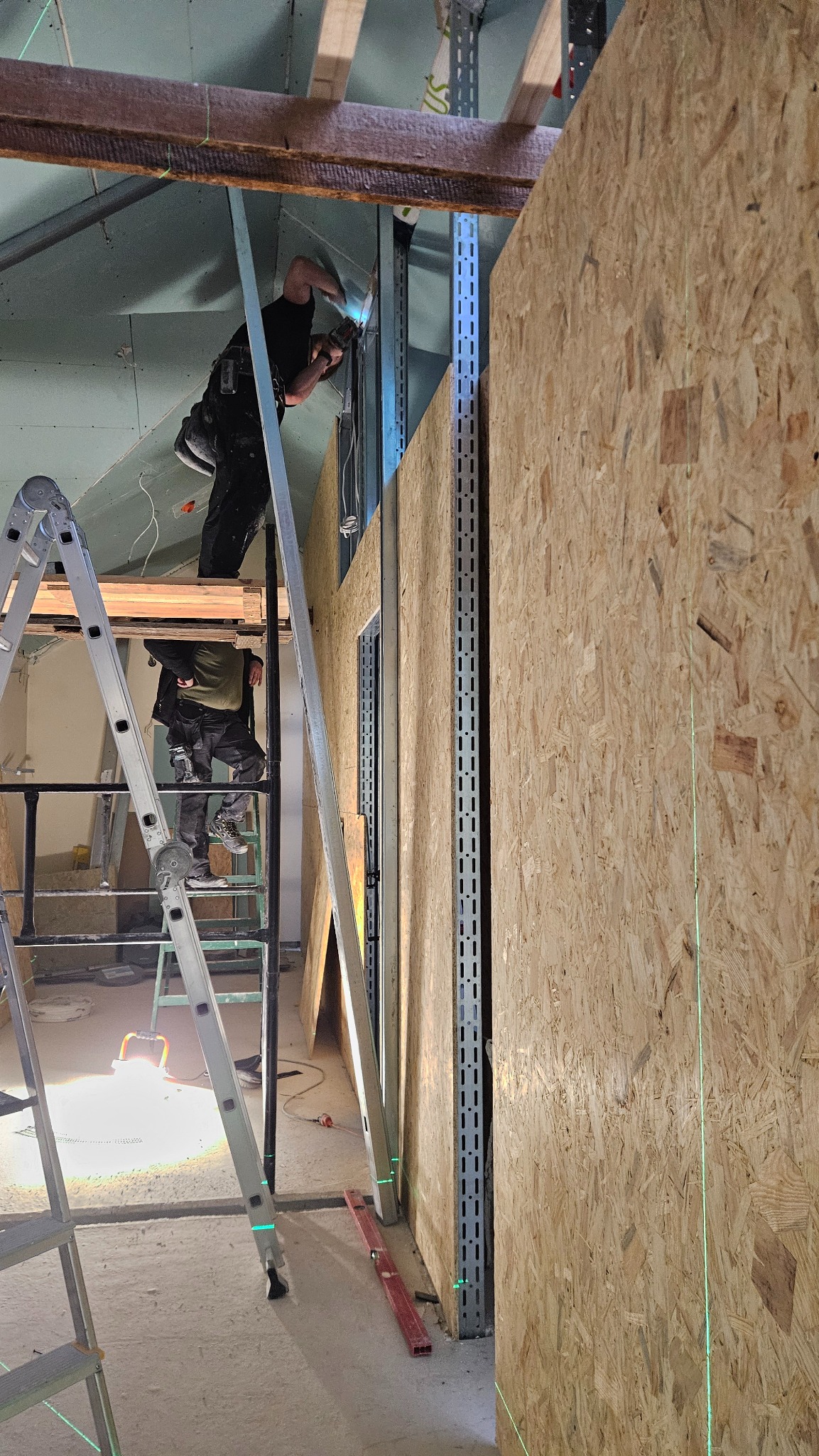 Montaż ścian z płyty OSB na metalowym stelażu. Widoczny pracownik na rusztowaniu, poziomica laserowa i fragment sufitu z płyt kartonowo-gipsowych. Prace wykończeniowe.