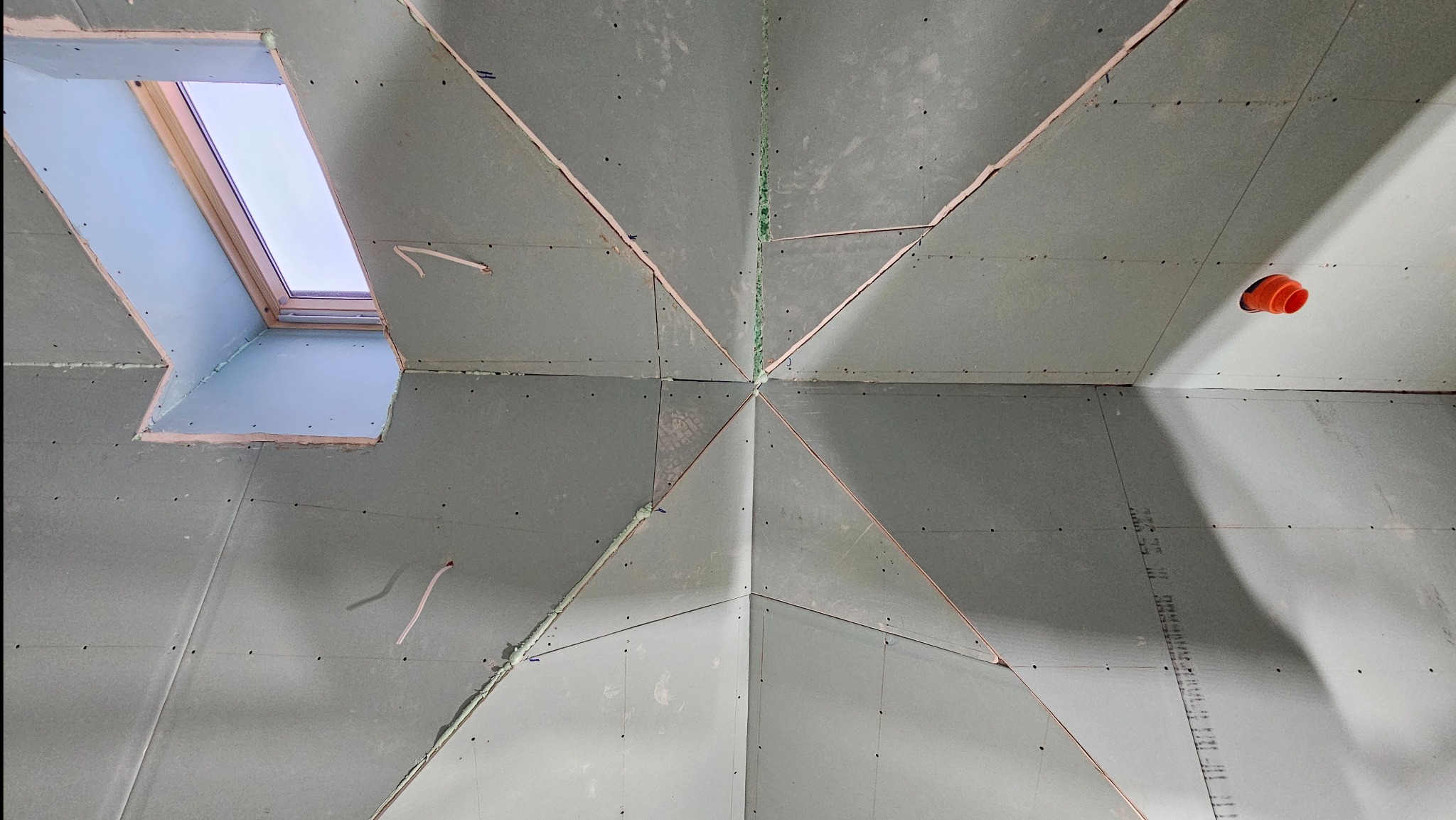 Poddasze z oknem dachowym w trakcie wykańczania płytami gipsowo-kartonowymi. Widoczne łączenia płyt i otwór wentylacyjny. Ujęcie z dołu, podkreślające geometrię.