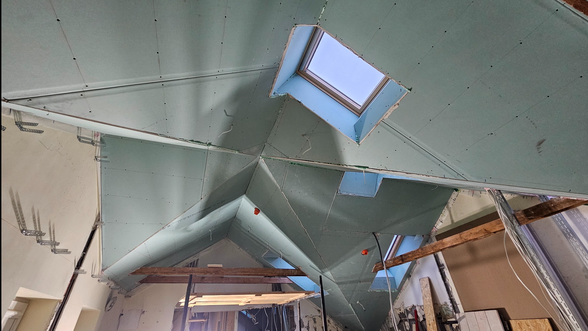 Skośny sufit poddasza z zamontowanymi płytami gipsowo-kartonowymi, widoczne okna dachowe i elementy konstrukcyjne. Prace wykończeniowe w toku.