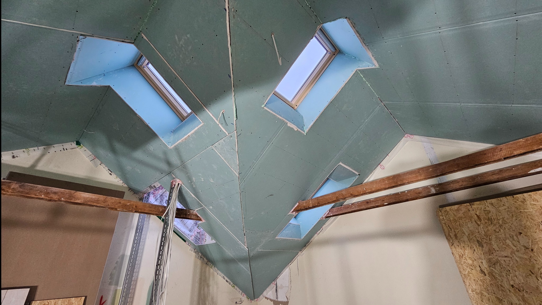 Skośny sufit z płyt gipsowo-kartonowych z oknami dachowymi, widok z dołu. Widoczne drewniane belki i fragmenty ścian. Etap prac wykończeniowych.